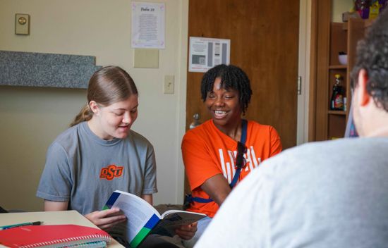 Two girls sitting on a bed laughing as they read a book.